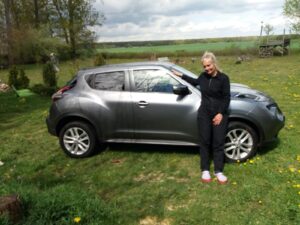 silbernes Auto steht auf einer Wiese, Frau steht davor und hält die Hand auf das Auto